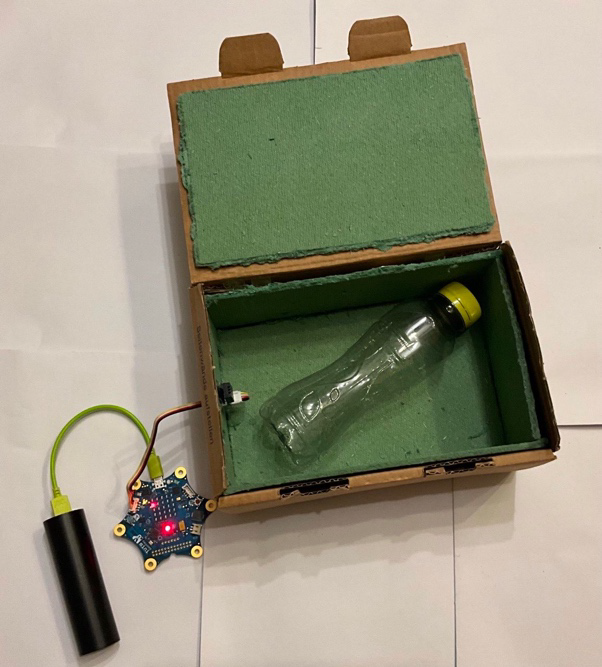 Messung der Temperatur mit Calliope mini und externen Sensoren: Abgebildet ist ein Pappkarton, der an den Innenflächen mit einem grünen Material gedämmt ist. Im Inneren des Kartons liegt eine Wasserflasche als Wärmequelle. Ein Temperatursensor misst dort die Temperatur. Er ist durch eine kleine Öffnung an einen Calliope mini außerhalb angeschlossen.